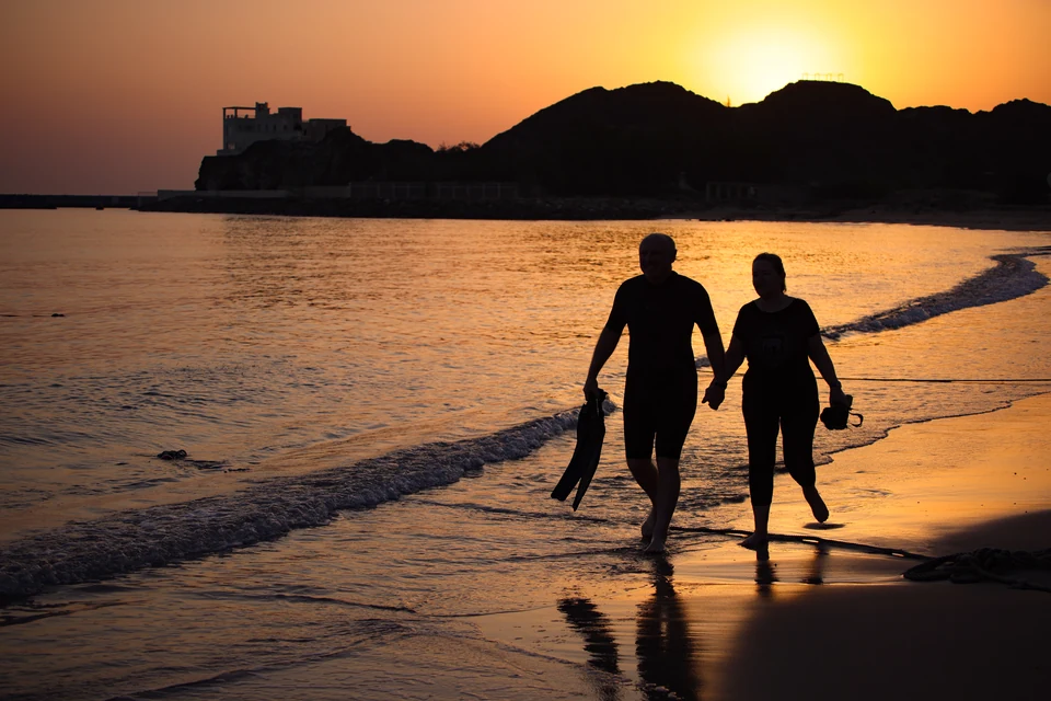 Two guests walking along the shoreline carrying snorkeling equipment in golden light.