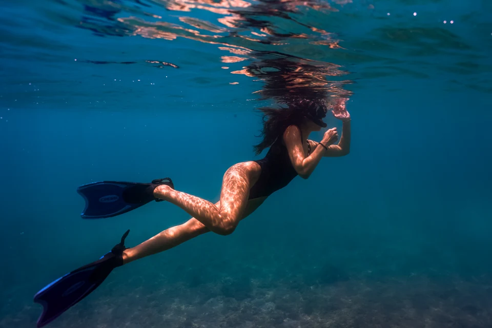 A woman in a black swimsuit and blue fins snorkeling beneath the water’s surface.